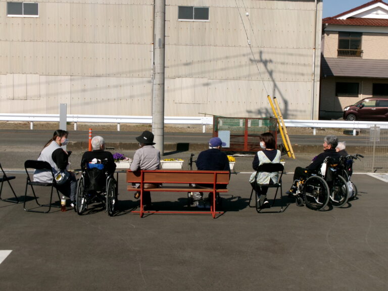 施設の屋外スペースで入居者が車いすに乗って集まり、テーブルを囲んで交流している様子